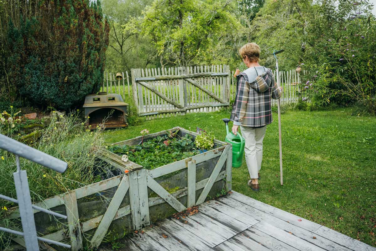 Die Hausbesitzerin läuft mit Gießkanne und Rechen in der Hand durch den Garten ihres alten Hauses.