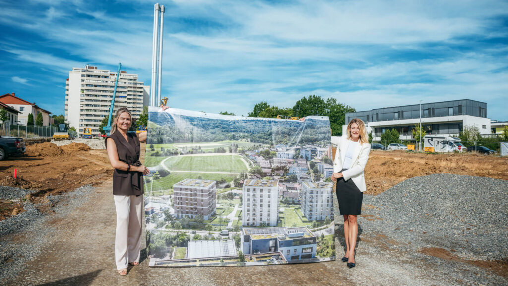 Zwei Maklerinnen halten auf der Baustelle ein großes Plakat mit einer Ansicht der drei geplanten Gebäude.
