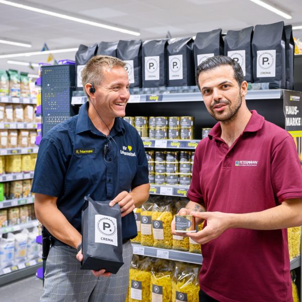 Ronald Petermann und sein Mitarbeiter George präsentieren lächelnd Produkte der regionalen Hausmarke P.