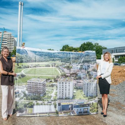 Zwei Maklerinnen halten auf der Baustelle ein großes Plakat mit einer Ansicht der drei geplanten Gebäude.