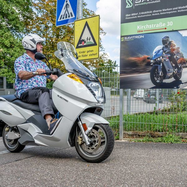Michael fährt mit dem E-Roller und schaut dabei auf ein Plakat, das ein E-Motorrad zeigt.
